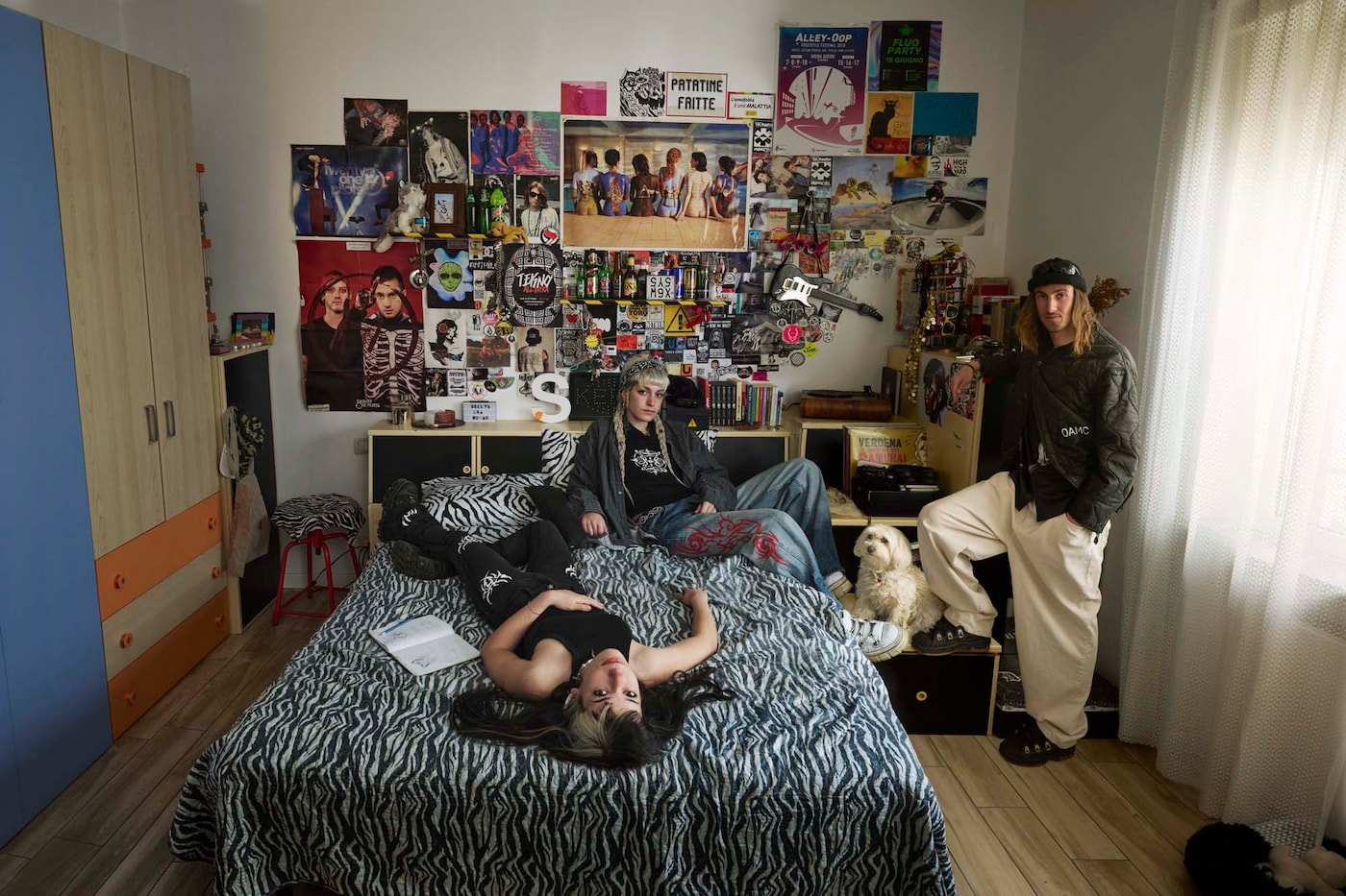 Three people lounging in a bedroom with a bed dressed in zebra stripe sheets and a wall covered of posters, art, stickers and souvenirs. 