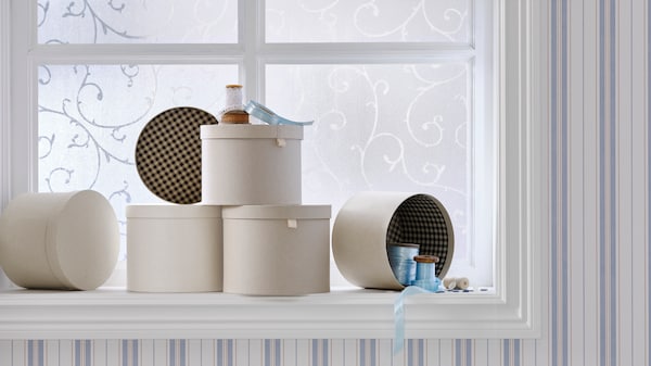 RÅGODLING storage boxes with lids are on a windowsill. One box is on its side, revealing the checked pattern inside.